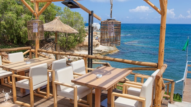 A wooden deck with white chairs and tables overlooking a beach with a clear blue sea. - Elmi Suites Beach Hotel - All Inclusive (Hersonissos)