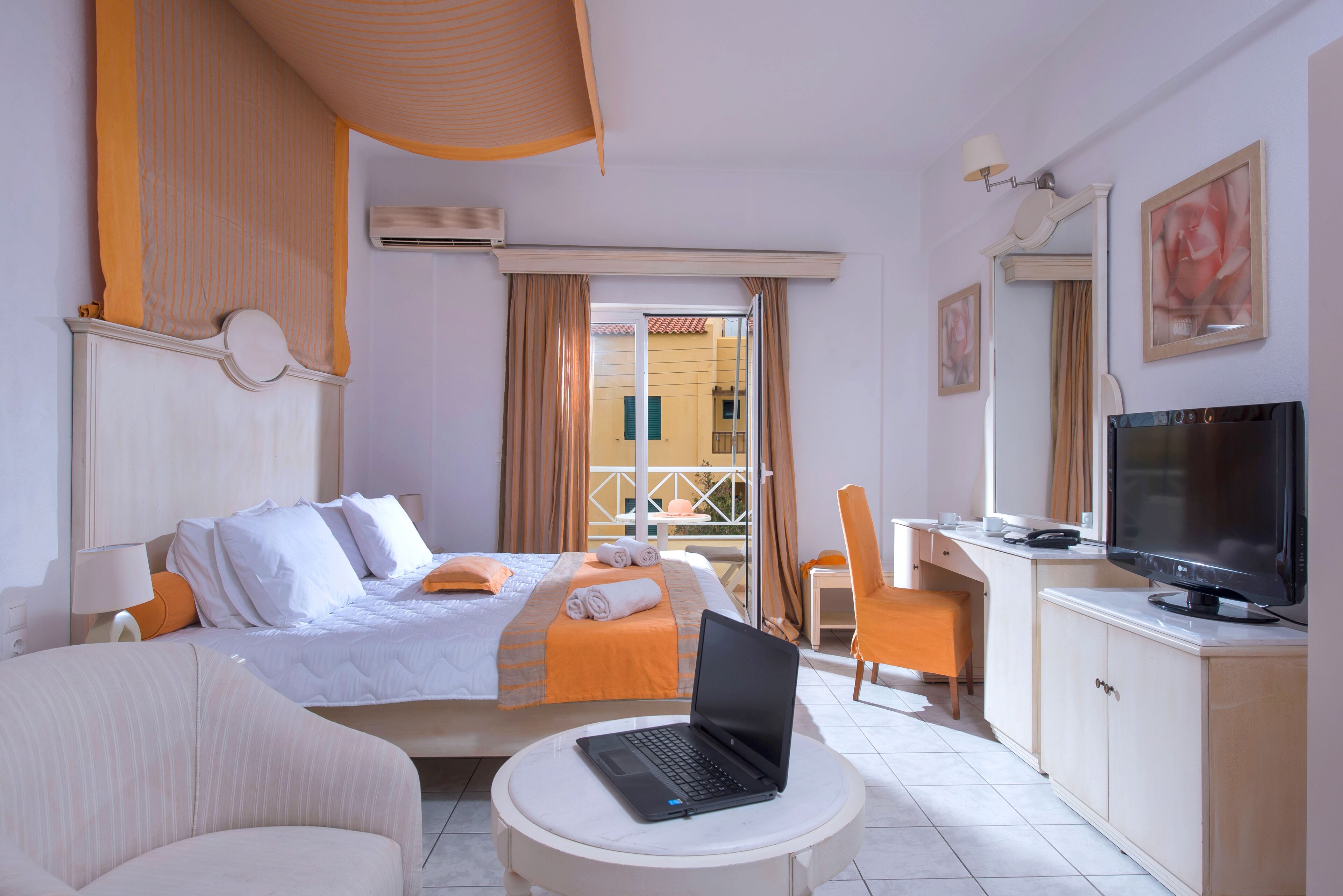 A hotel room with a bed, a desk with a laptop, a TV, a chair, and a window with curtains.