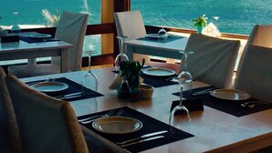 A dining table set with plates, cutlery, and wine glasses, overlooking a coastal view with a body of water and distant land. - Elmi Suites Beach Hotel - All Inclusive (Hersonissos)