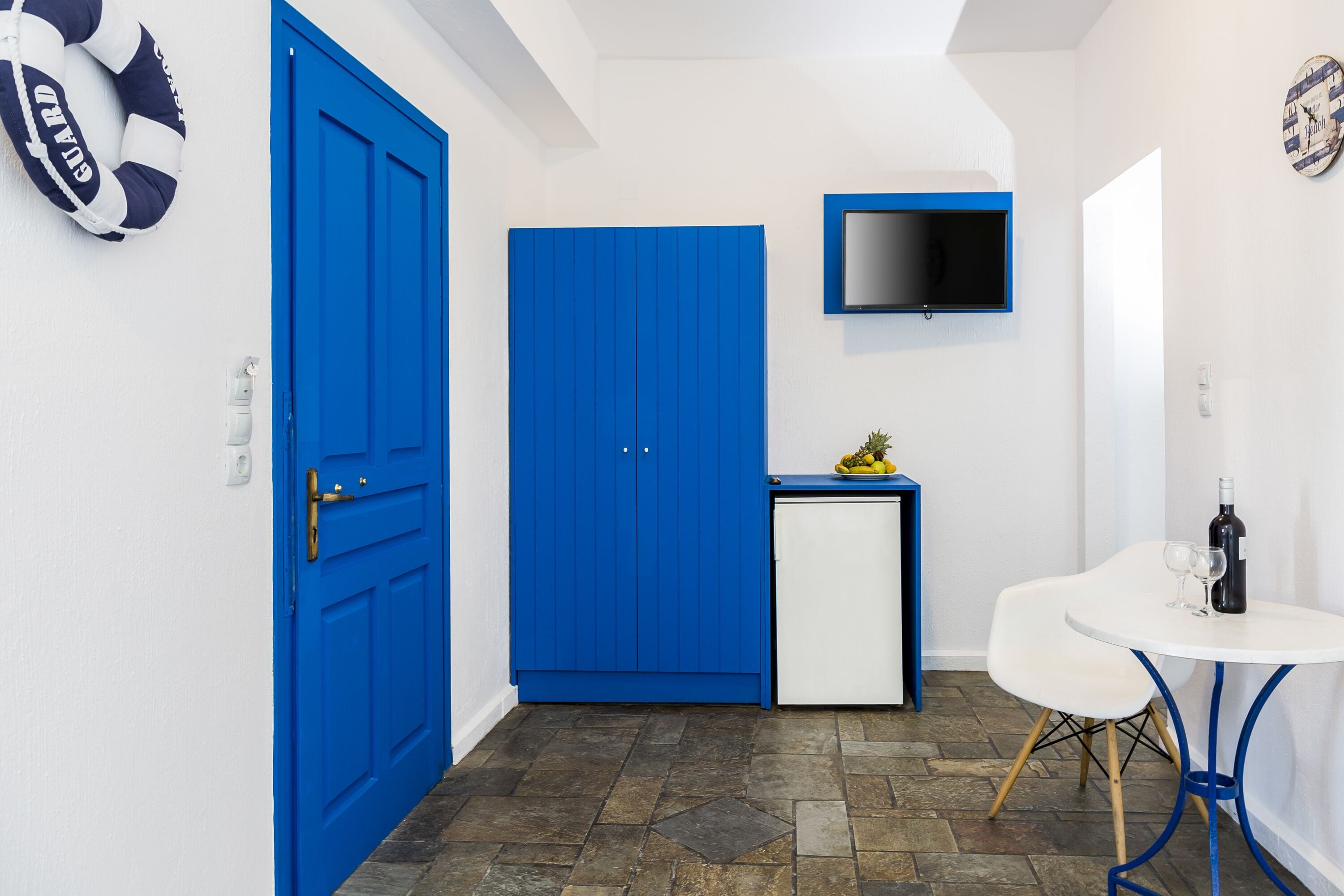 A compact room with a blue door, a small white fridge, a television mounted on the wall, and a white table with a bottle and glasses.