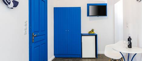 A compact room with a blue door, a small white fridge, a television mounted on the wall, and a white table with a bottle and glasses.