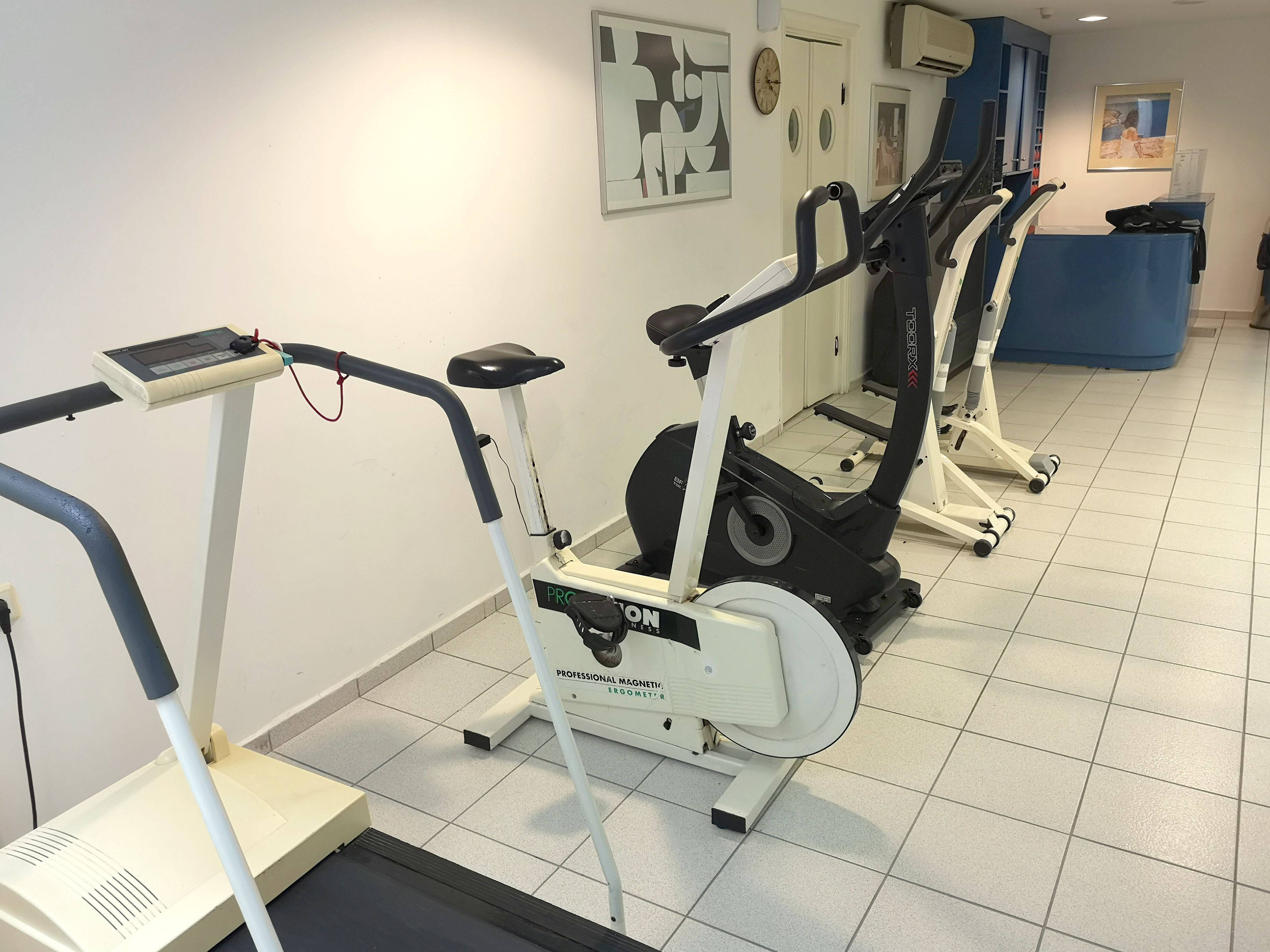 Un gimnasio con varias bicicletas estáticas y cintas de correr, un mostrador de recepción y un reloj de pared.