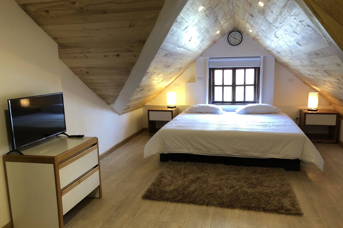 Une chambre avec un plafond en pente, un lit avec une literie blanche, deux lampes de chevet, un téléviseur posé sur un meuble et une commode en bois.