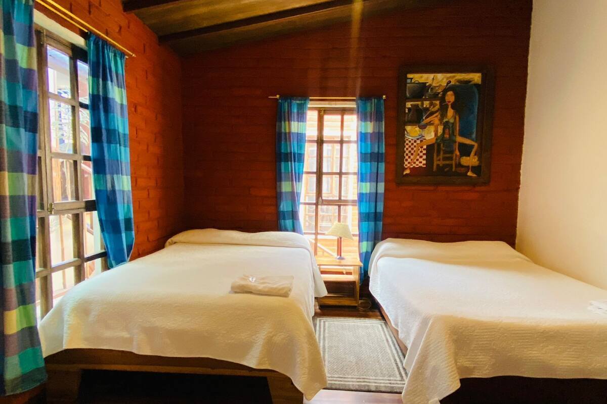 Une chambre avec deux lits, une fenêtre avec des rideaux, un tableau au mur et un plancher en bois.