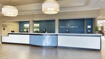 Uma receção de hotel com um posto de auto-check-in digital, zona de serviços empresariais e uma grande parede azul com o nome do hotel.