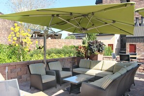 Un patio exterior con mobiliario de mimbre, un parasol verde y plantas en macetas.