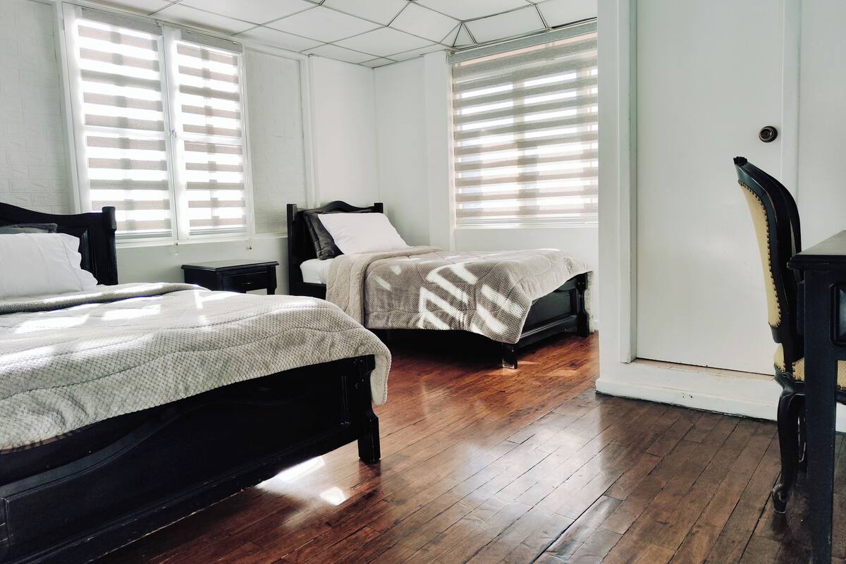 A bedroom with two beds, wooden flooring, and large windows with blinds.