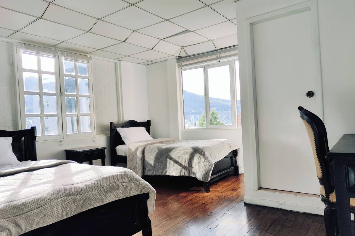 A room with two beds, a chair, a desk, and large windows with a city view.