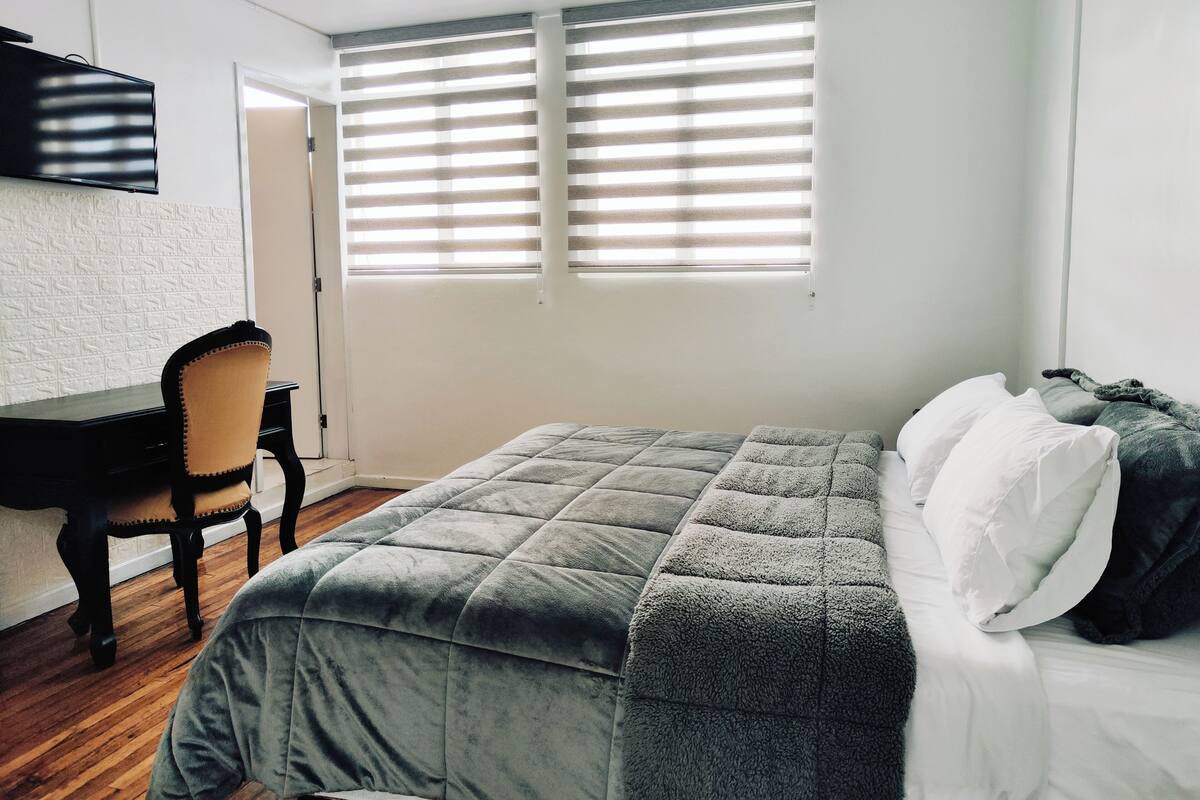 A bedroom with a bed, a desk, and a chair. There is a window with blinds.