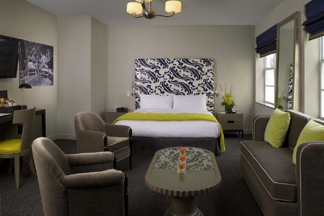 Une chambre d’hôtel comprenant un lit, deux fauteuils, un canapé, une petite table, un bureau, une chaise, un téléviseur, une fenêtre avec des rideaux et une œuvre d’art accrochée au mur.