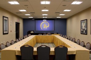 A conference room with a long table, chairs, a projector screen, and a nameplate.