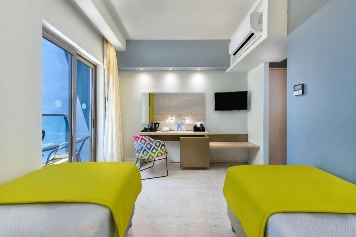 A hotel room with a balcony, a desk with a chair, and a bed with bright yellow covers.
