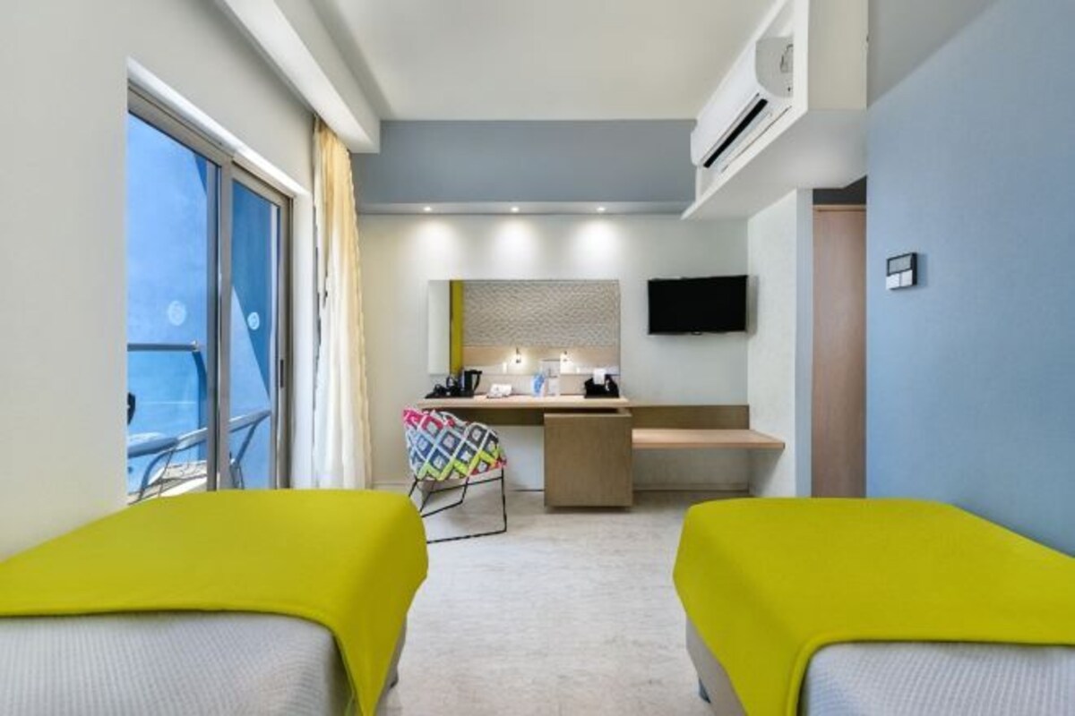 Quarto de hotel com duas cadeiras amarelas, uma escrivaninha com cadeira, uma cama, televisão e janela com vista.
