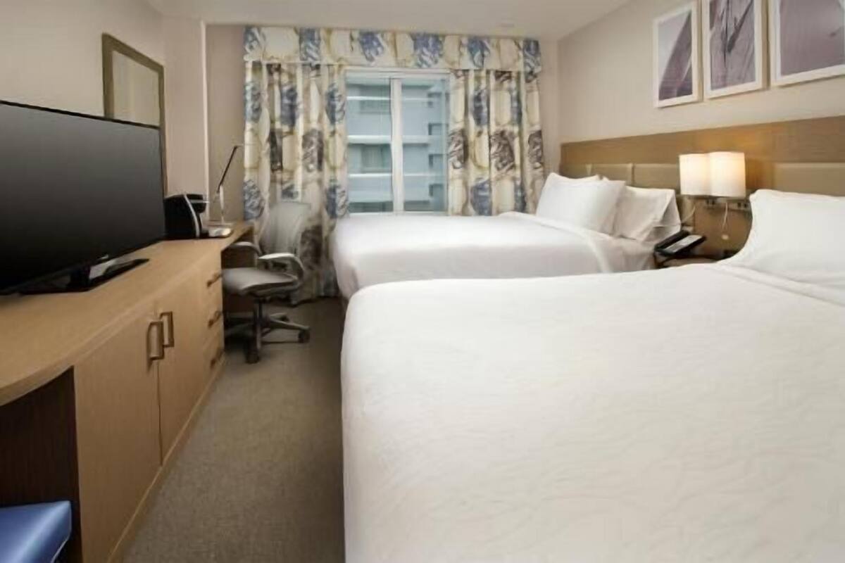 Une chambre d’hôtel avec deux lits, un bureau avec une chaise, une télévision et une fenêtre avec des rideaux.