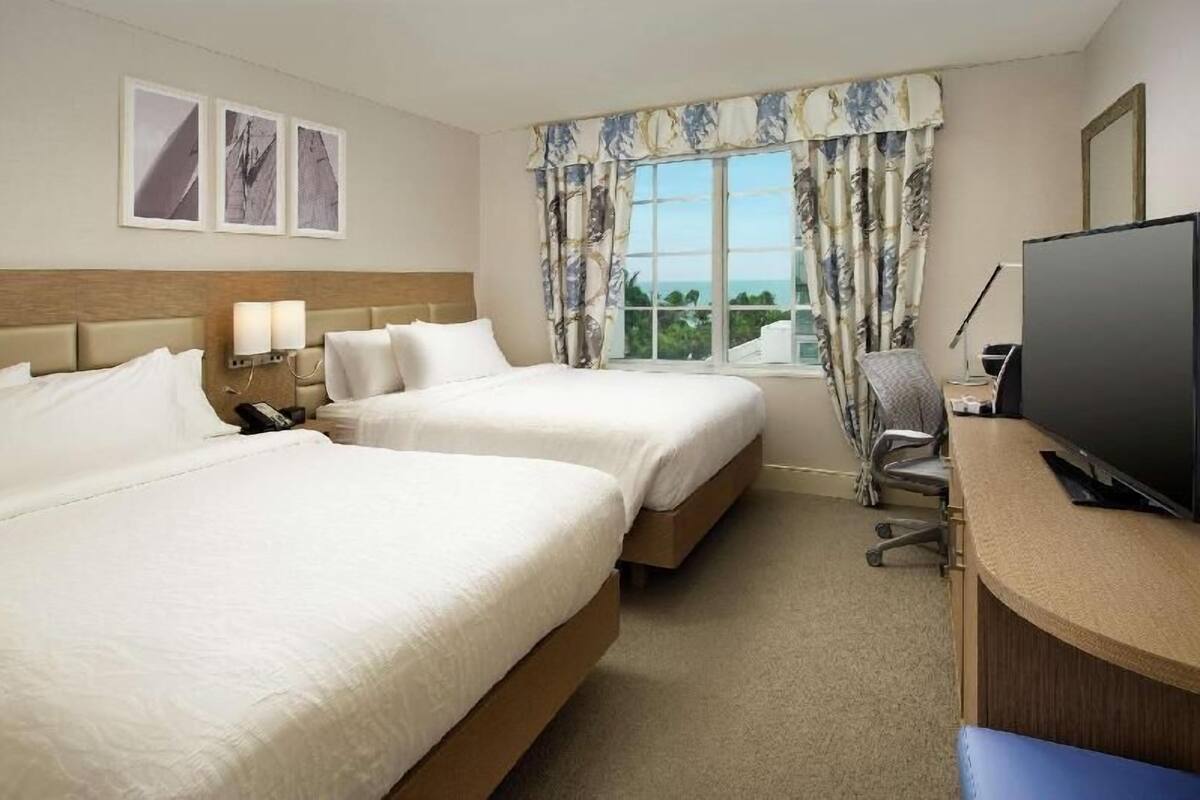 Une chambre d’hôtel avec deux lits, un bureau avec une télévision, une chaise et une fenêtre avec des rideaux.