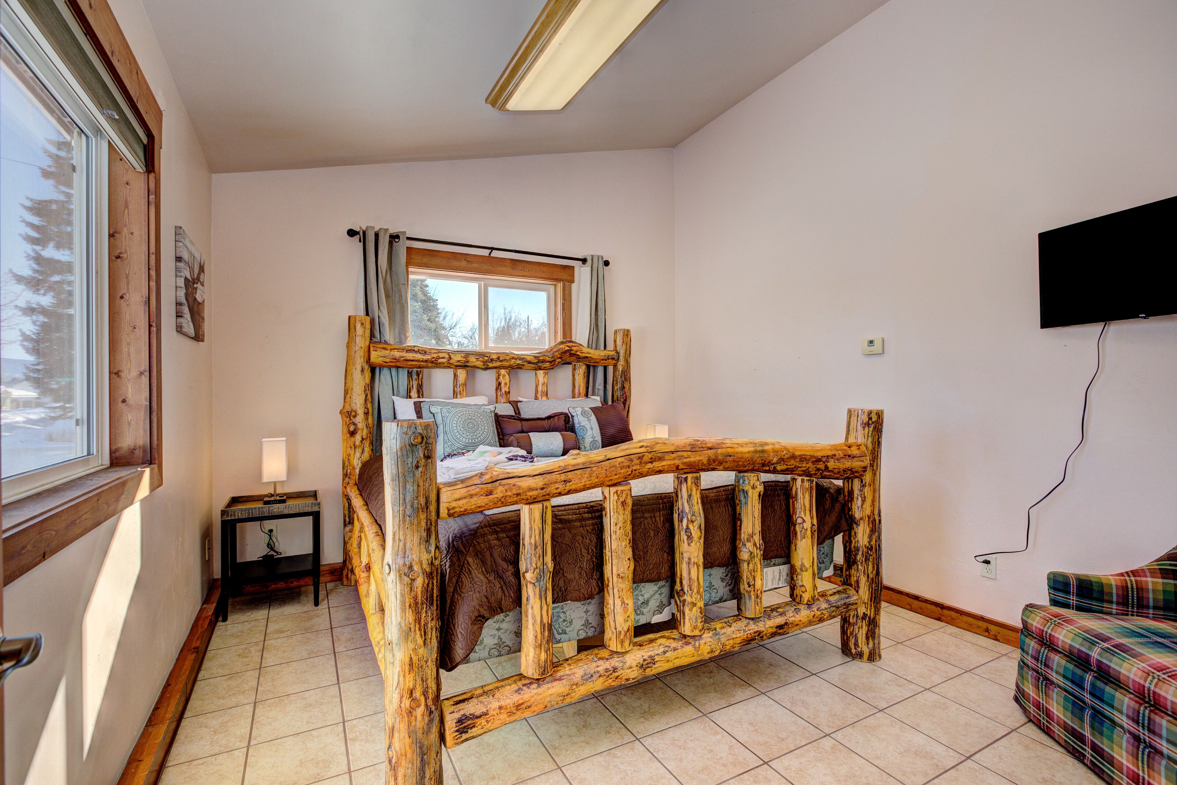 A room with a wooden bunk bed, a small table with a lamp, a window with curtains, and a television mounted on the wall.
