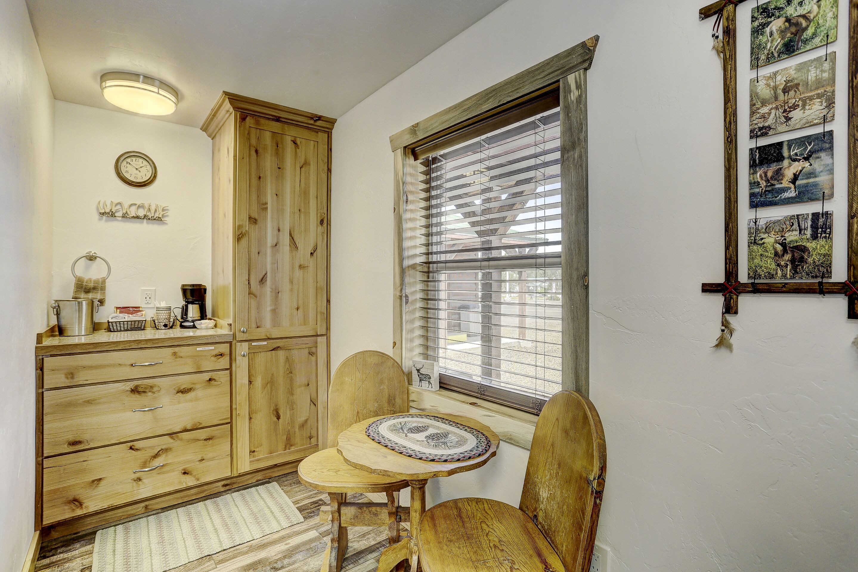 A room with a wooden cabinet, a small table with chairs, a window with blinds, and a wall clock.