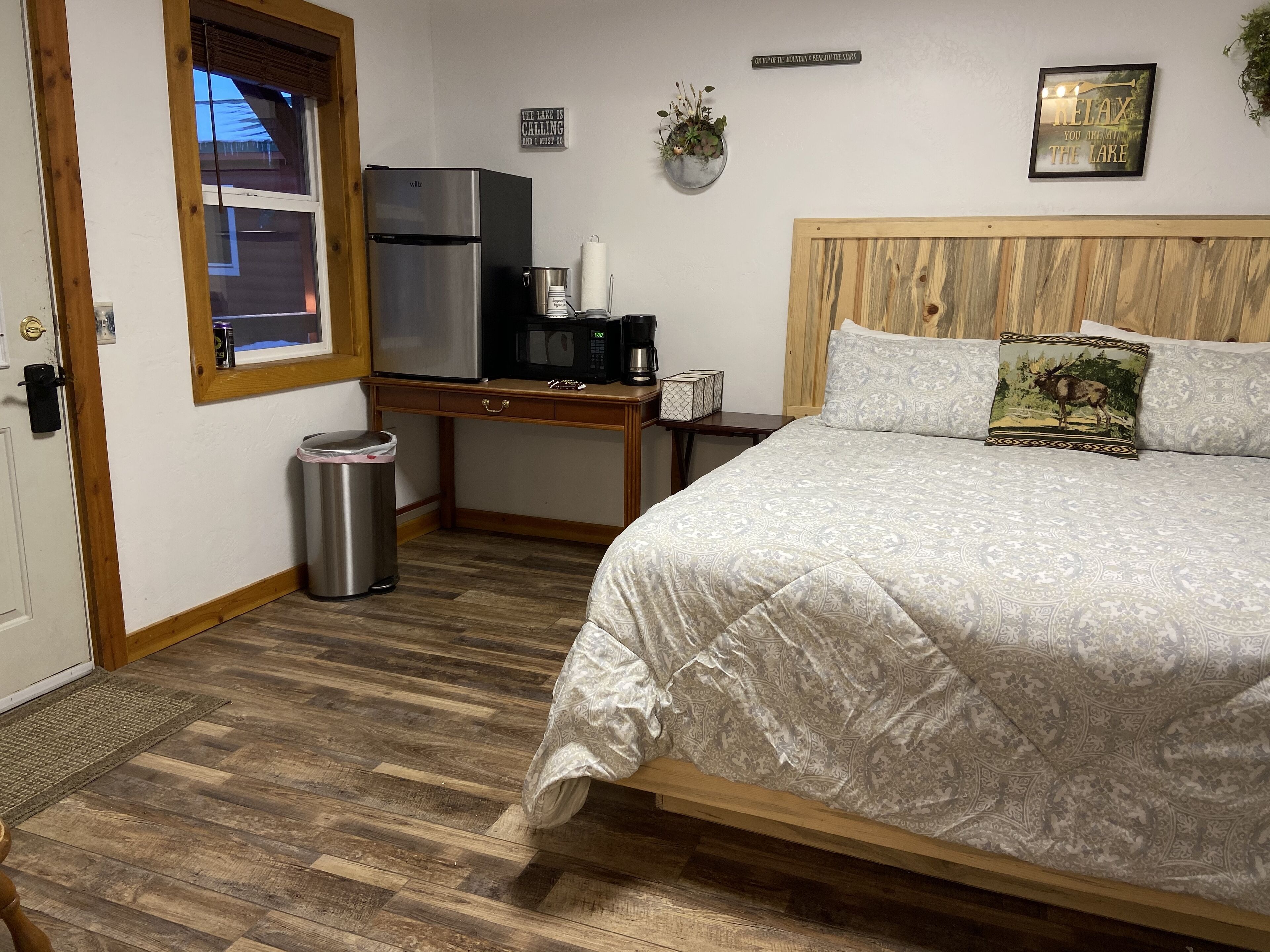 A bedroom with a bed, wooden headboard, a small desk with a refrigerator, microwave, and coffee maker. There is a window with blinds, a trash can, and a wall-mounted sign.