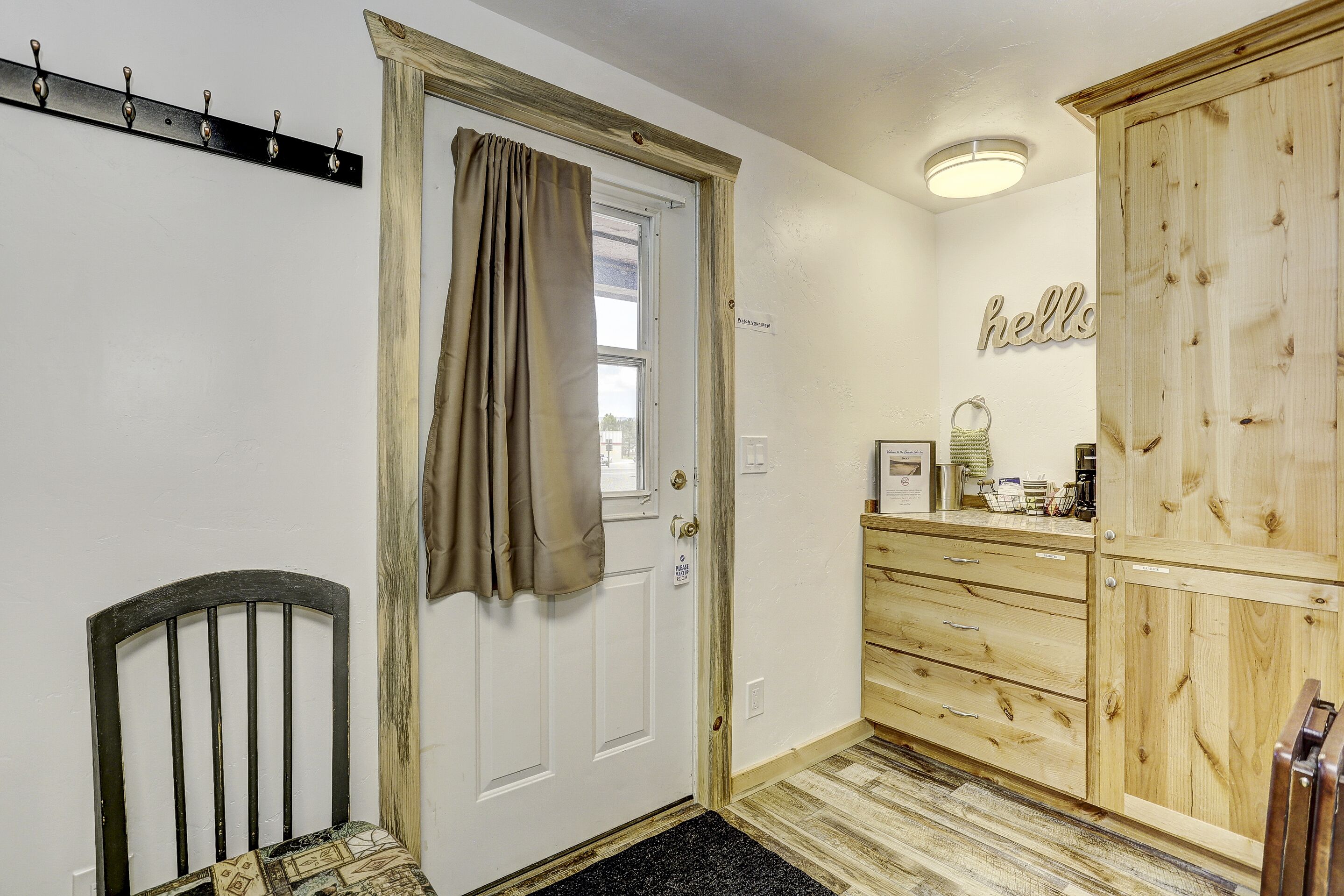 Un couloir avec une porte blanche, un porte-manteau, une armoire en bois et une pancarte de bienvenue.