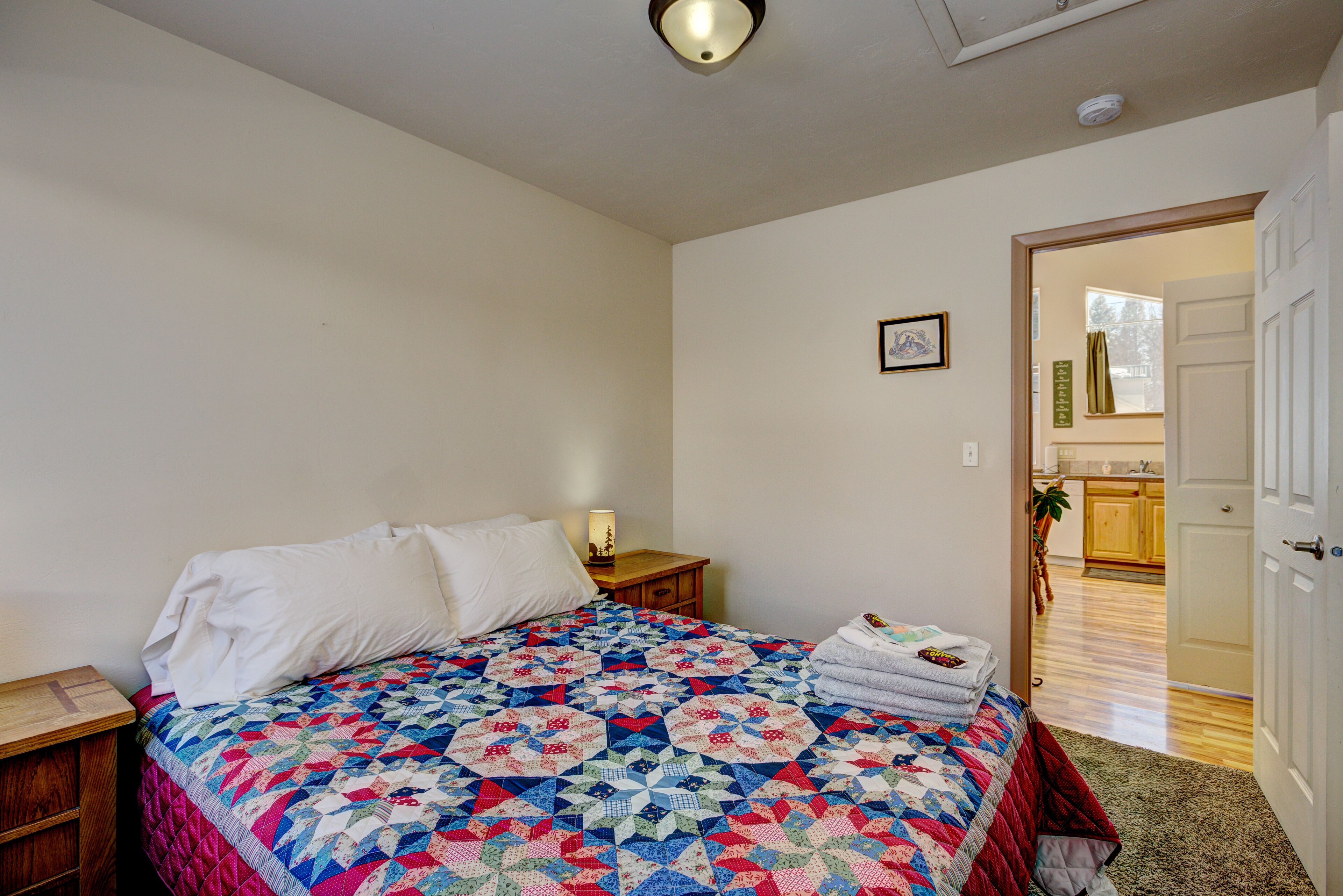 A bedroom with a large bed, a nightstand, a door leading to a kitchen, and a framed picture on the wall.