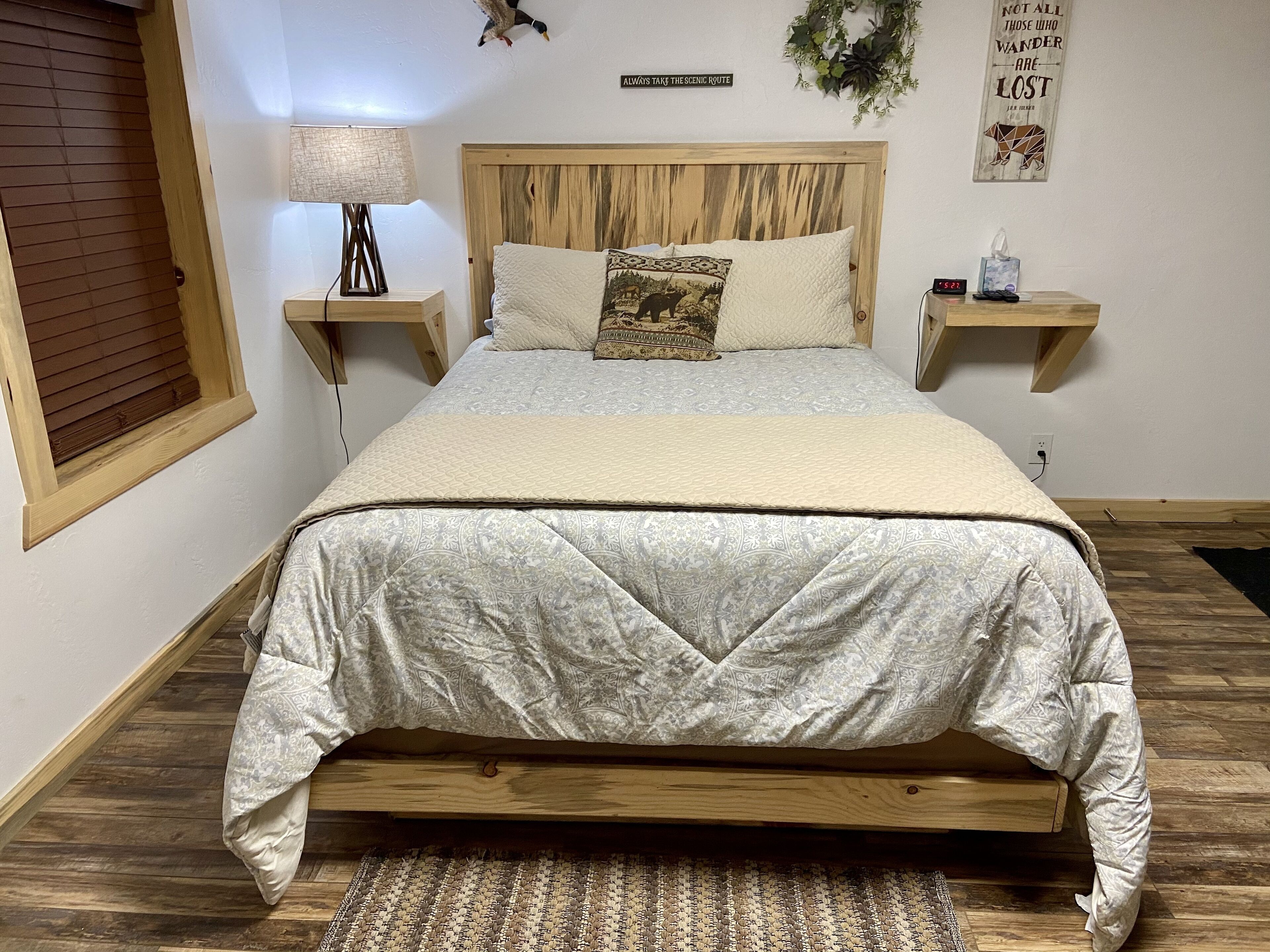 Ein Schlafzimmer mit einem hölzernen Bettgestell, einem gemusterten Überwurf, einer Nachttischlampe, einer Uhr und einem motivierenden Zitat an der Wand.