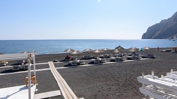 A beach with lounge chairs and umbrellas, a wooden deck, and a rocky cliff in the background. - Kamari Blue Boutique Hotel (Santorini)