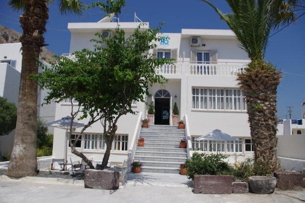 Un bâtiment blanc avec un escalier menant à l’entrée, entouré de palmiers et de plantes en pot.