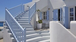 A white building with a staircase leading to a balcony, a potted plant, and a view of the sea. - Petinos Beach (Mykonos)