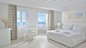 A bright, white room with a bed, a small table, and chairs, a view of the sea through a door, and a ceiling with wooden beams. - Petinos Beach (Mykonos)