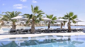 A poolside area with lounge chairs and umbrellas, palm trees, and a view of the sea and buildings in the background. - Petinos Beach (Mykonos)