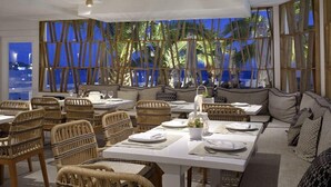 A modern dining area with wicker furniture, white tables, and a view of palm trees at night. - Petinos Beach (Mykonos)