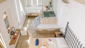 A hotel room with a bed, a small table with toiletries, a bathtub, and a window. - Antinea Suites And Spa (Santorini)
