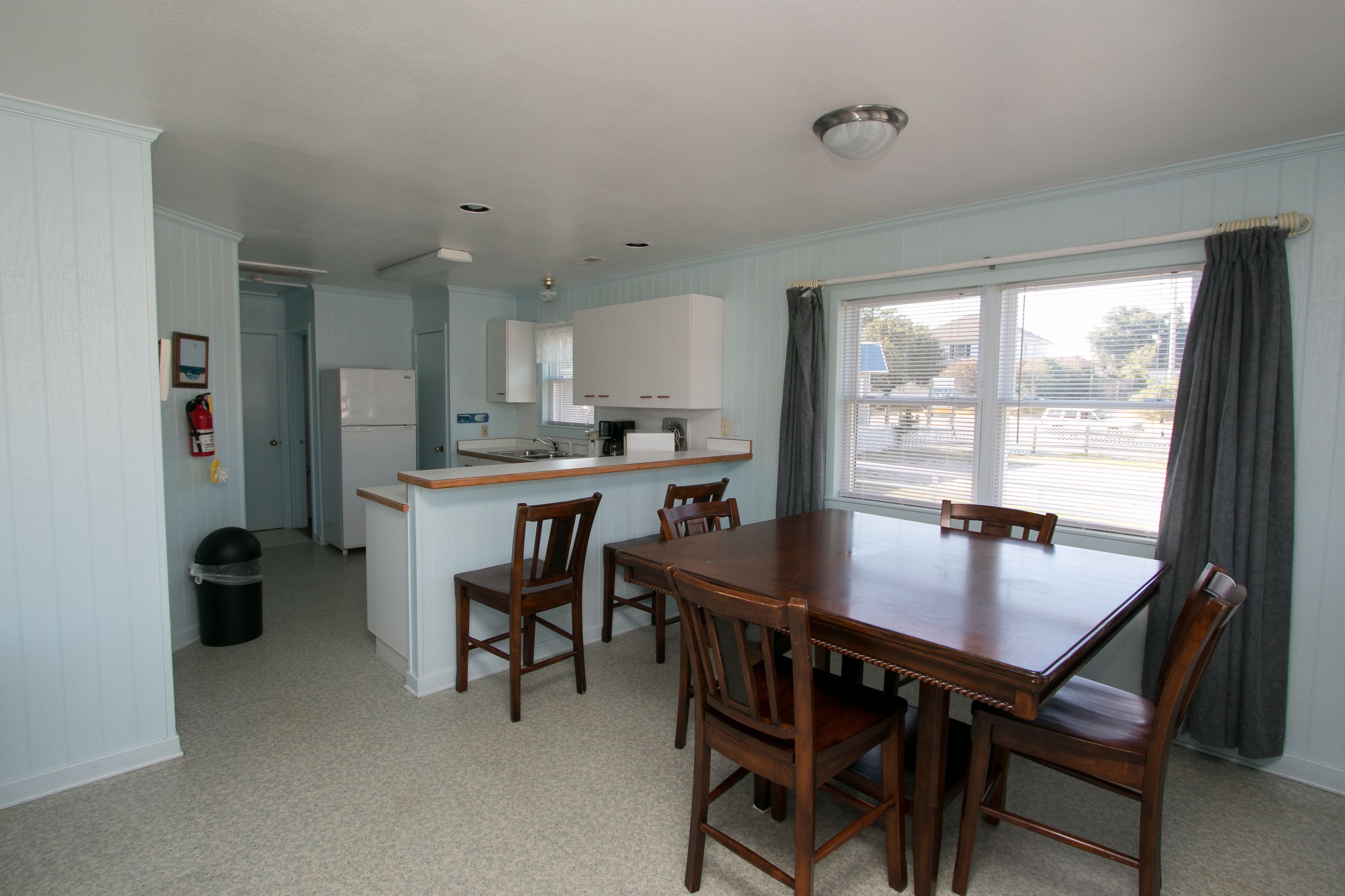 A dining area with a wooden table and chairs, a kitchen with white cabinets, and a window with curtains.