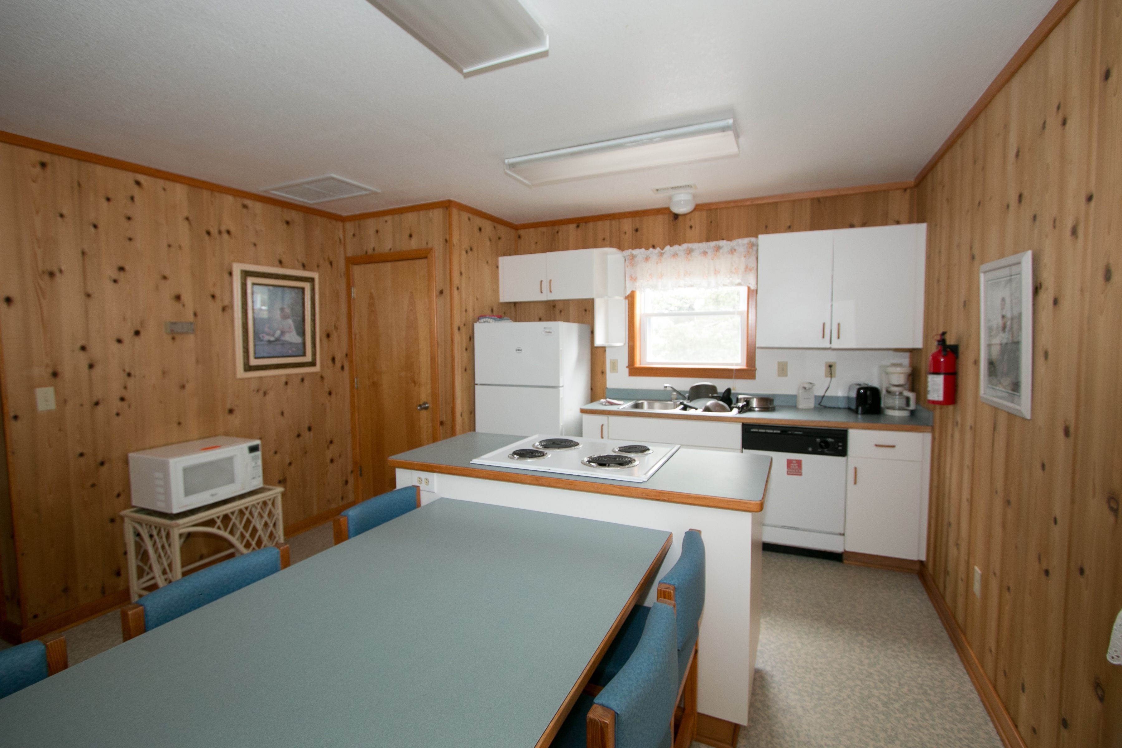 A compact kitchen with wooden paneling, a dining table with blue chairs, a microwave, a stove, and white cabinets.