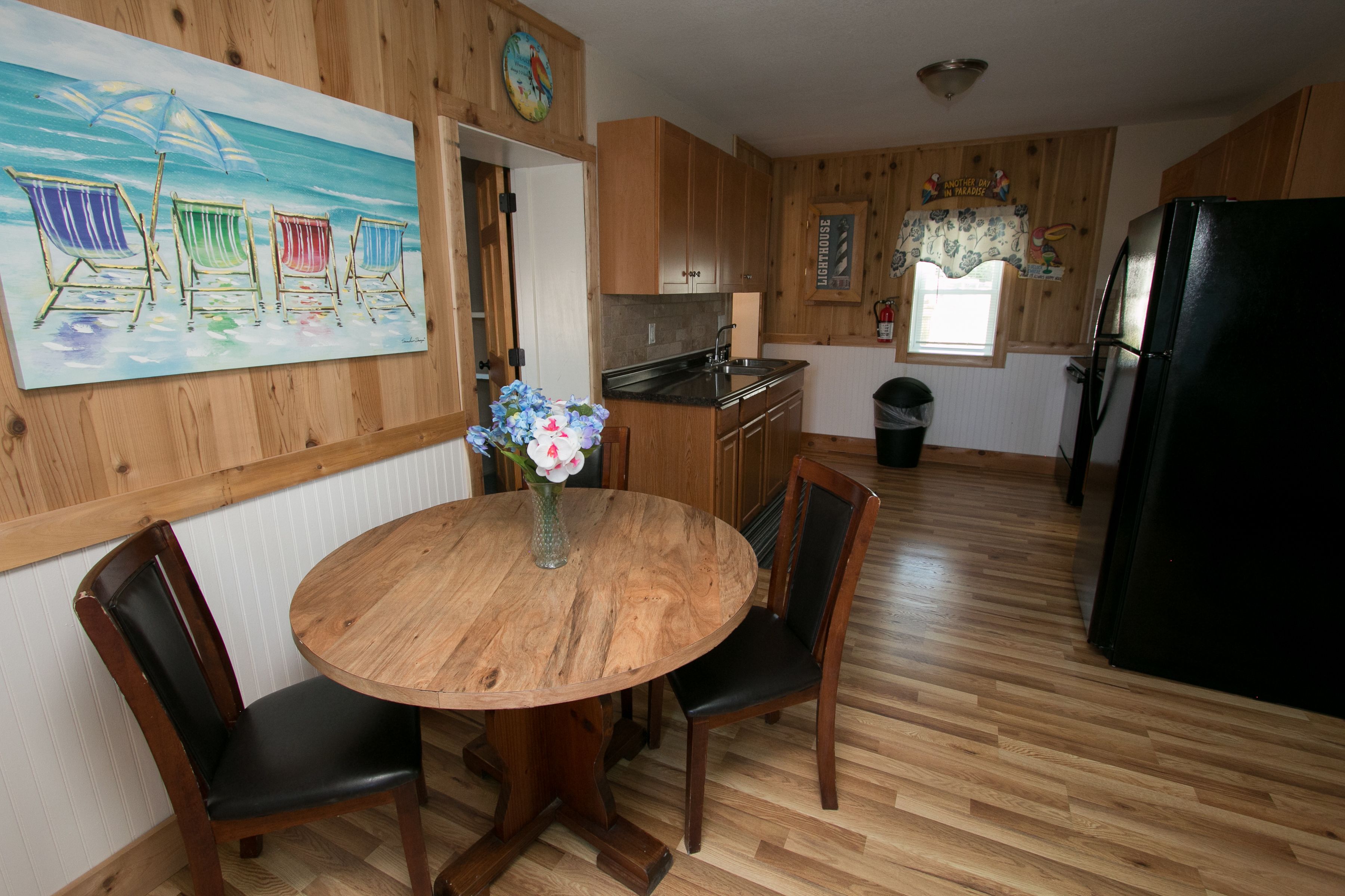 A wooden interior with a round dining table, chairs, a kitchen area, and a painting of beach chairs.