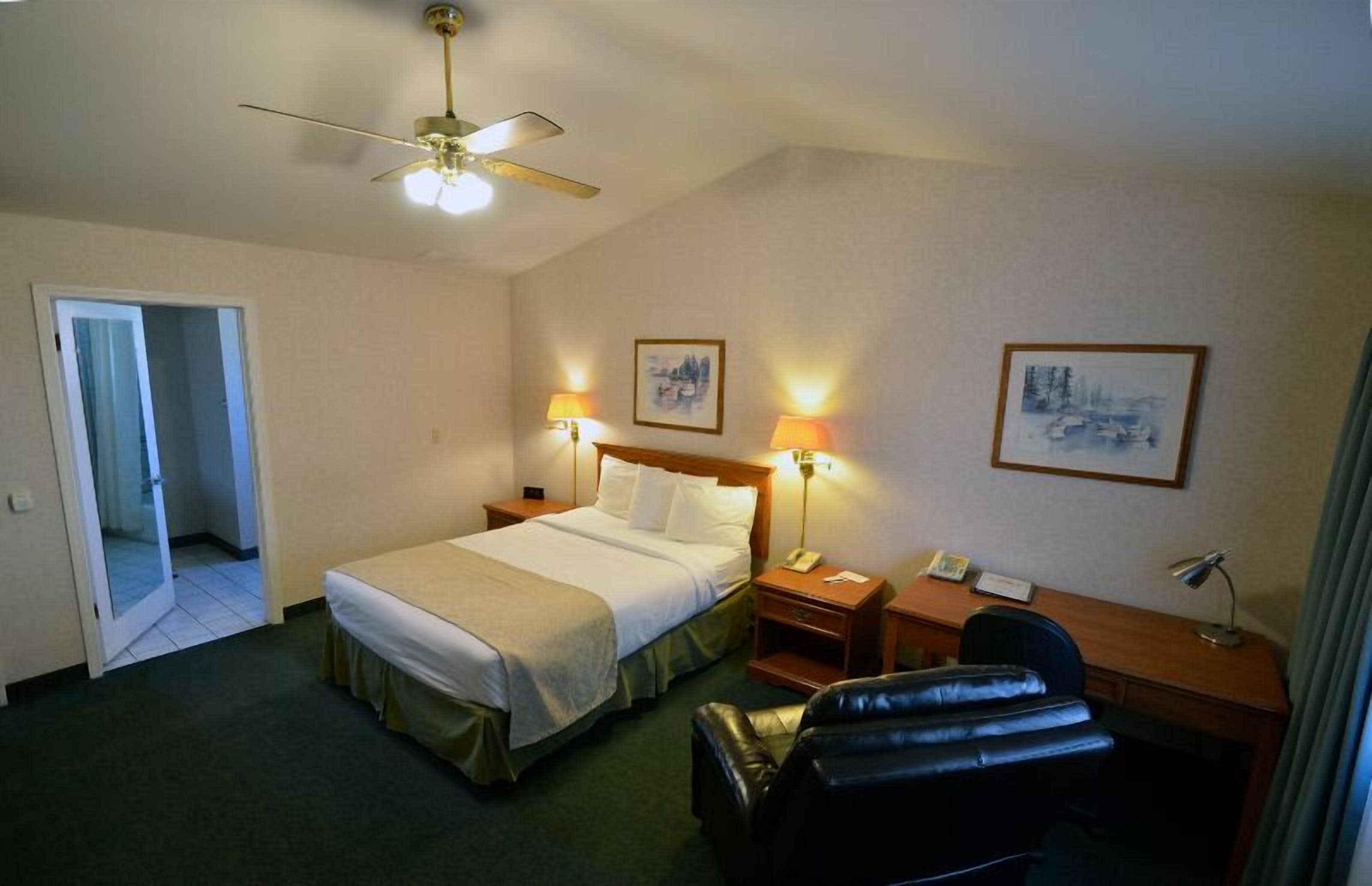A hotel room with a bed, bedside lamps, a desk, a chair, a ceiling fan, and two framed pictures on the wall.