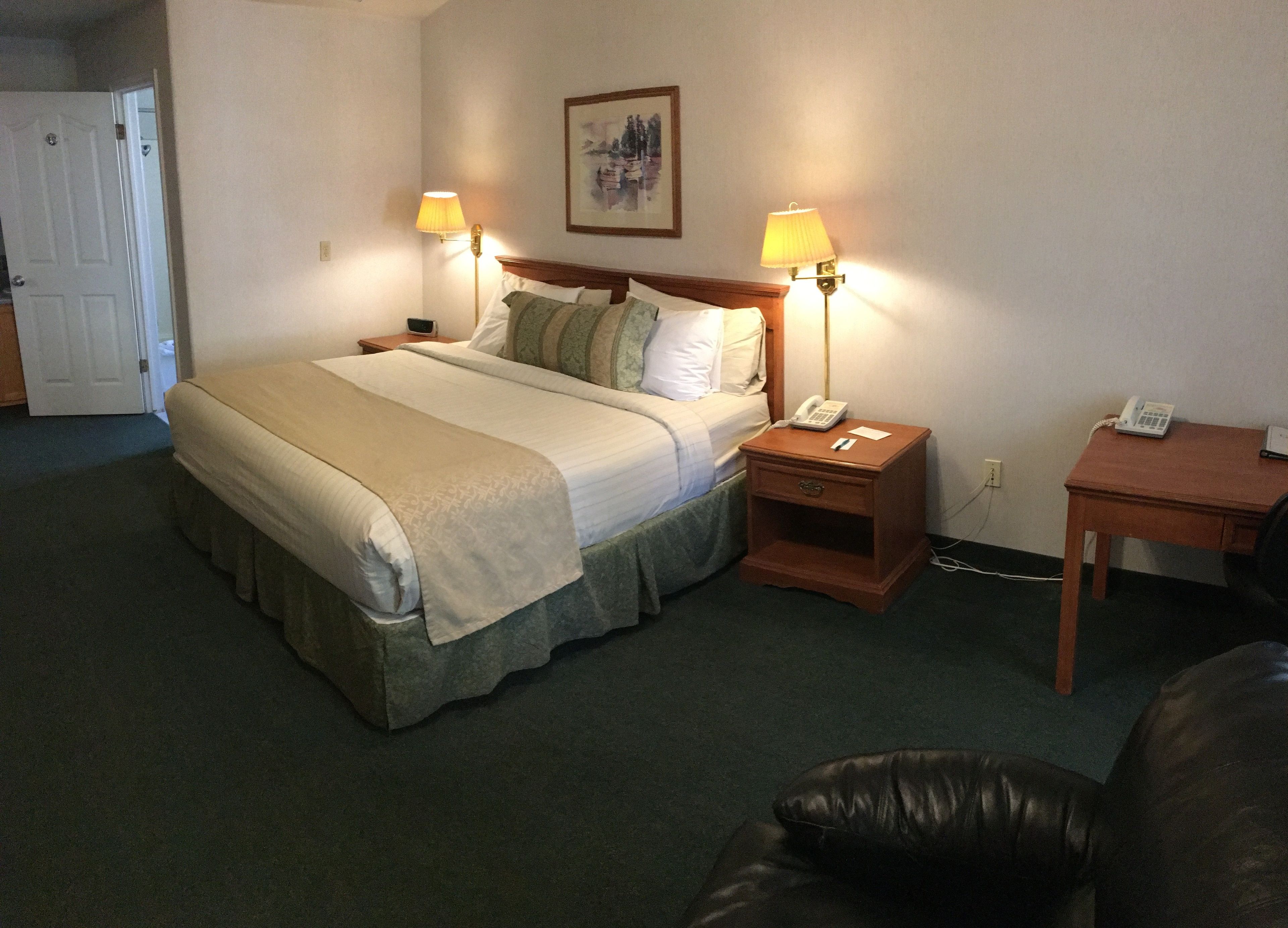 Quarto de hotel com uma cama, duas luminárias de cabeceira, mesa de cabeceira com telefone, escrivaninha com telefone e uma porta.