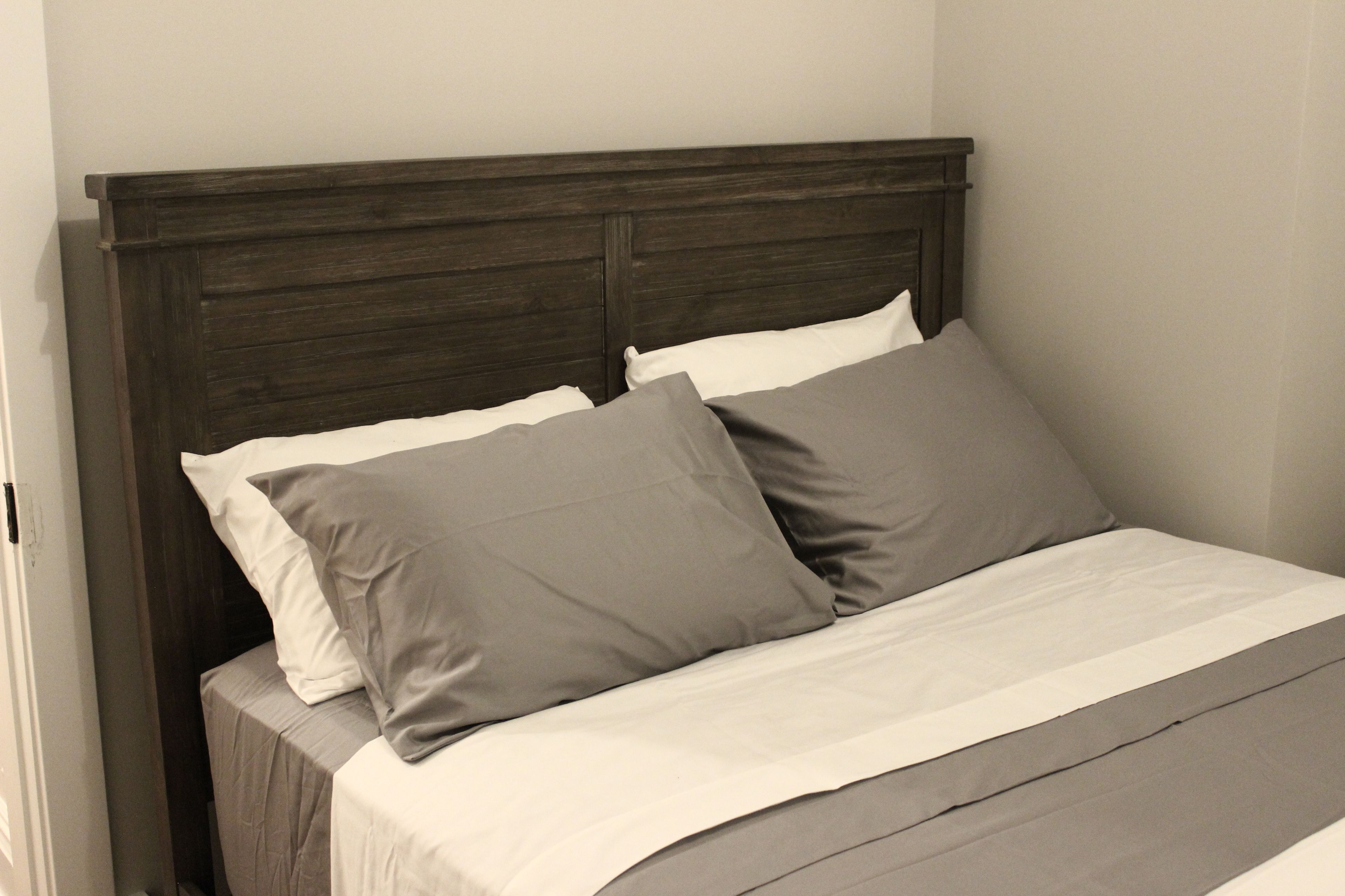 A bed with a dark wooden headboard, white and grey bedding, and a plain wall in the background.