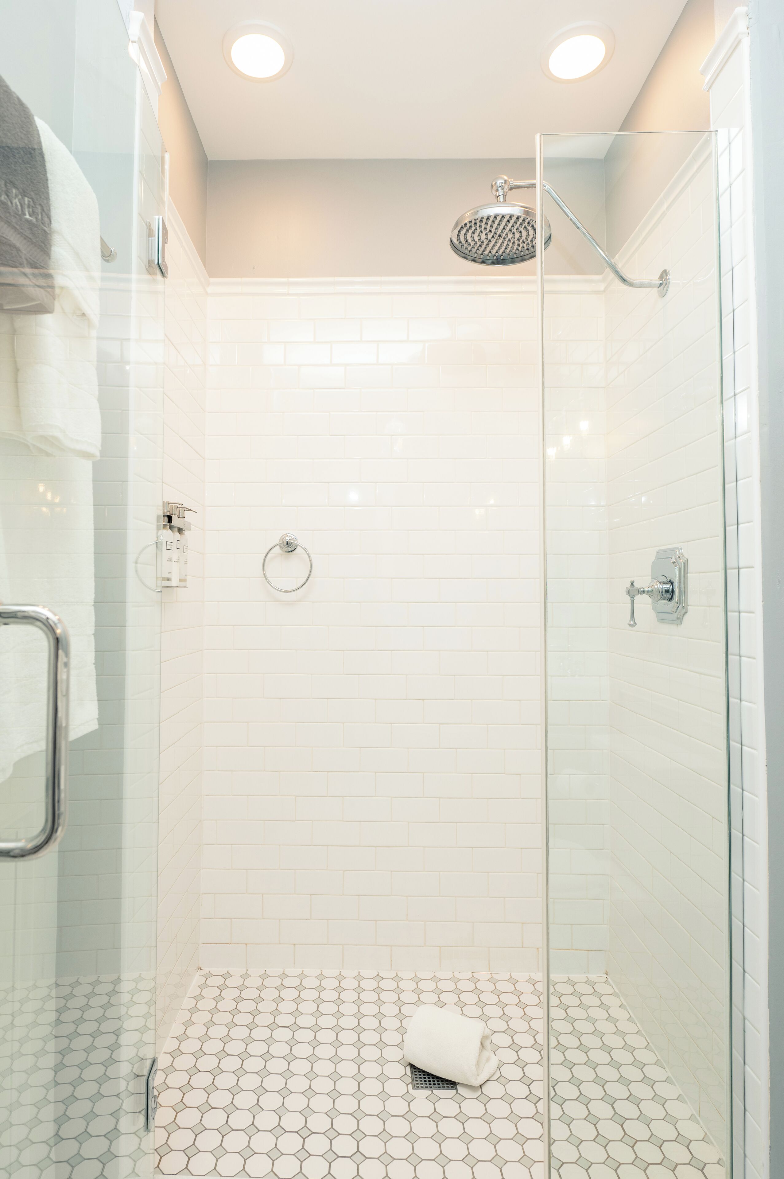 A glass-enclosed shower with white hexagonal tiles, a glass door, and a towel on the floor.