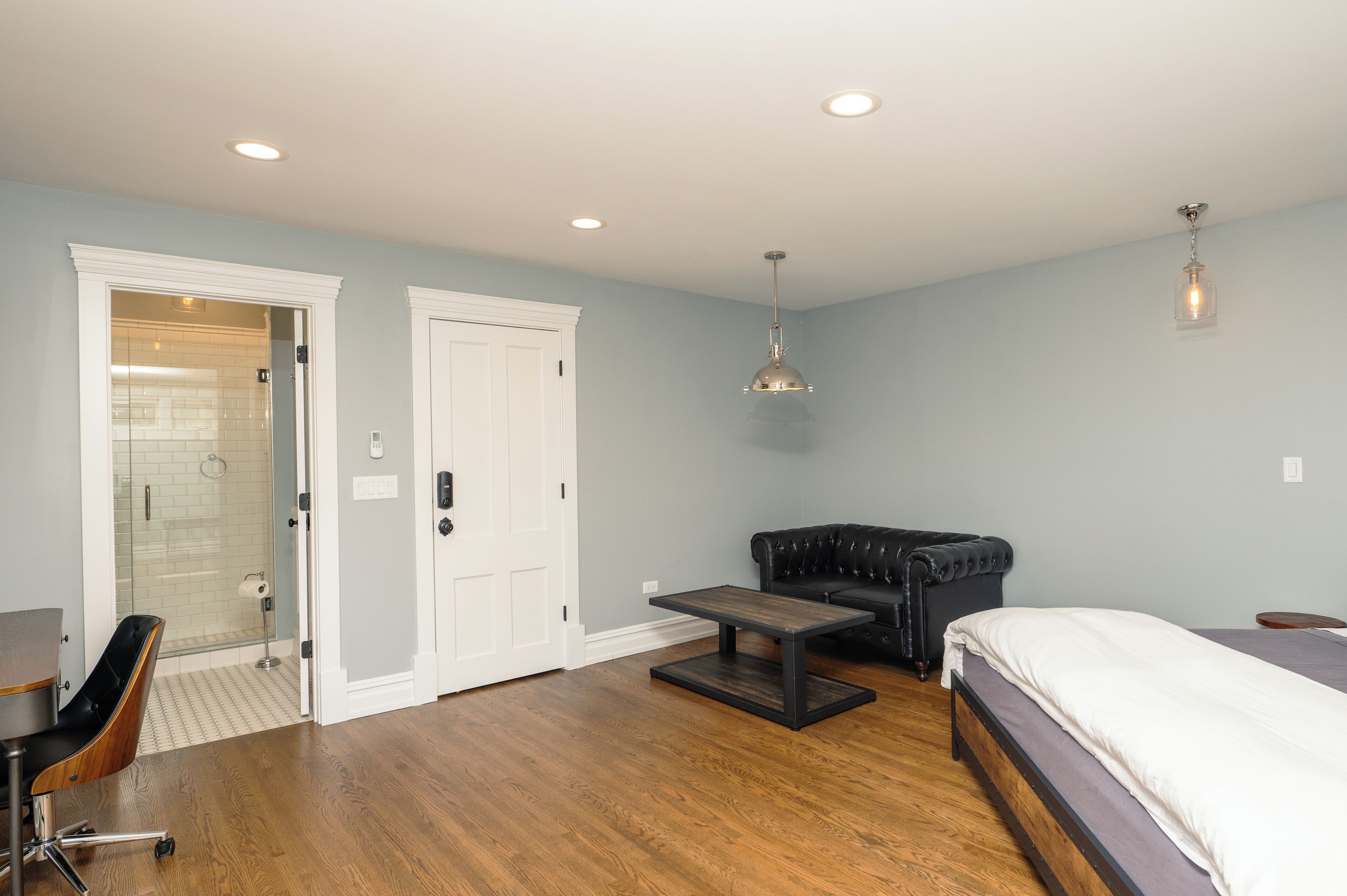 A bedroom with a bed, a black leather sofa, a wooden coffee table, and a glass shower enclosure.