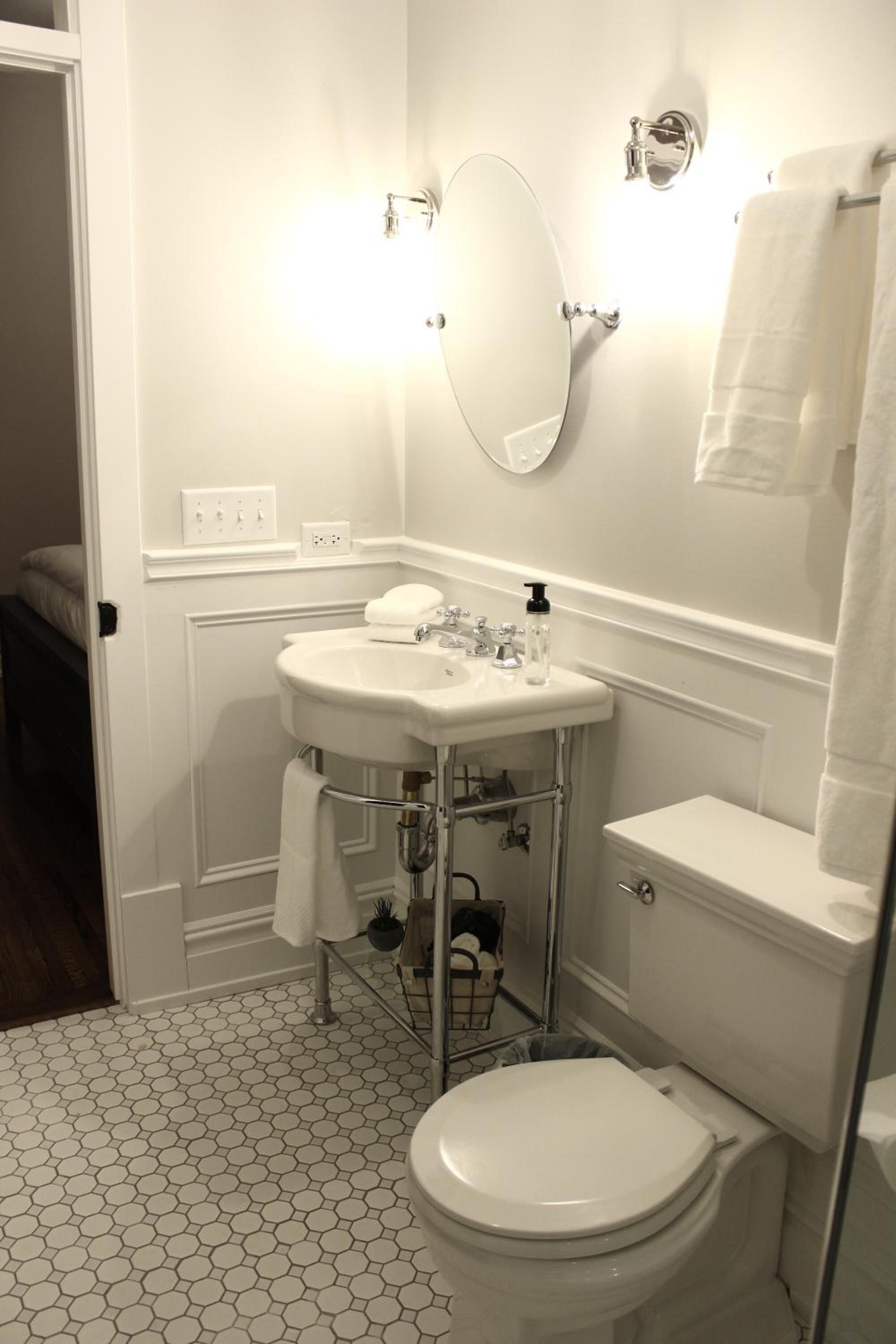 Une salle de bain avec un lavabo blanc, des toilettes et un miroir. On y trouve un porte-serviettes et un petit panier posé sur le sol.