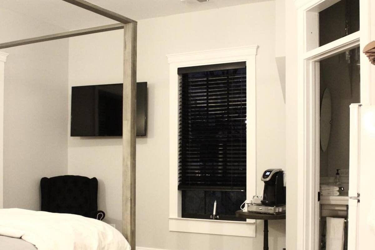 Une chambre à coucher avec un lit à baldaquin, un téléviseur fixé au mur, une fenêtre avec des stores, une machine à café et une salle de bain visible à travers une porte ouverte.