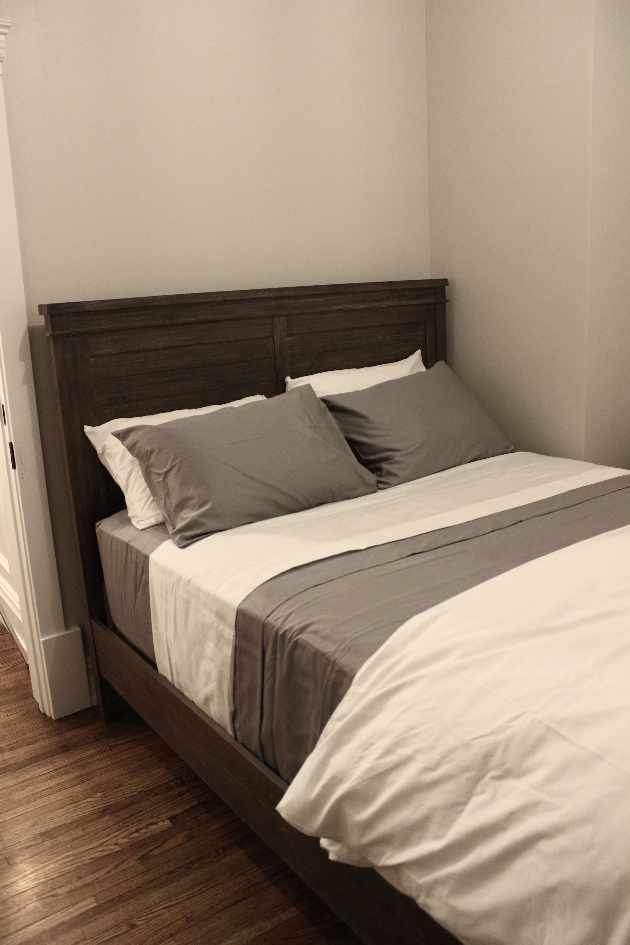 Una cama bien hecha con cabecera de madera, ropa de cama blanca y gris, y suelo de madera.