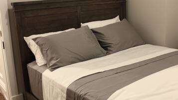 Una cama bien hecha con cabecera de madera, ropa de cama blanca y gris, y suelo de madera.