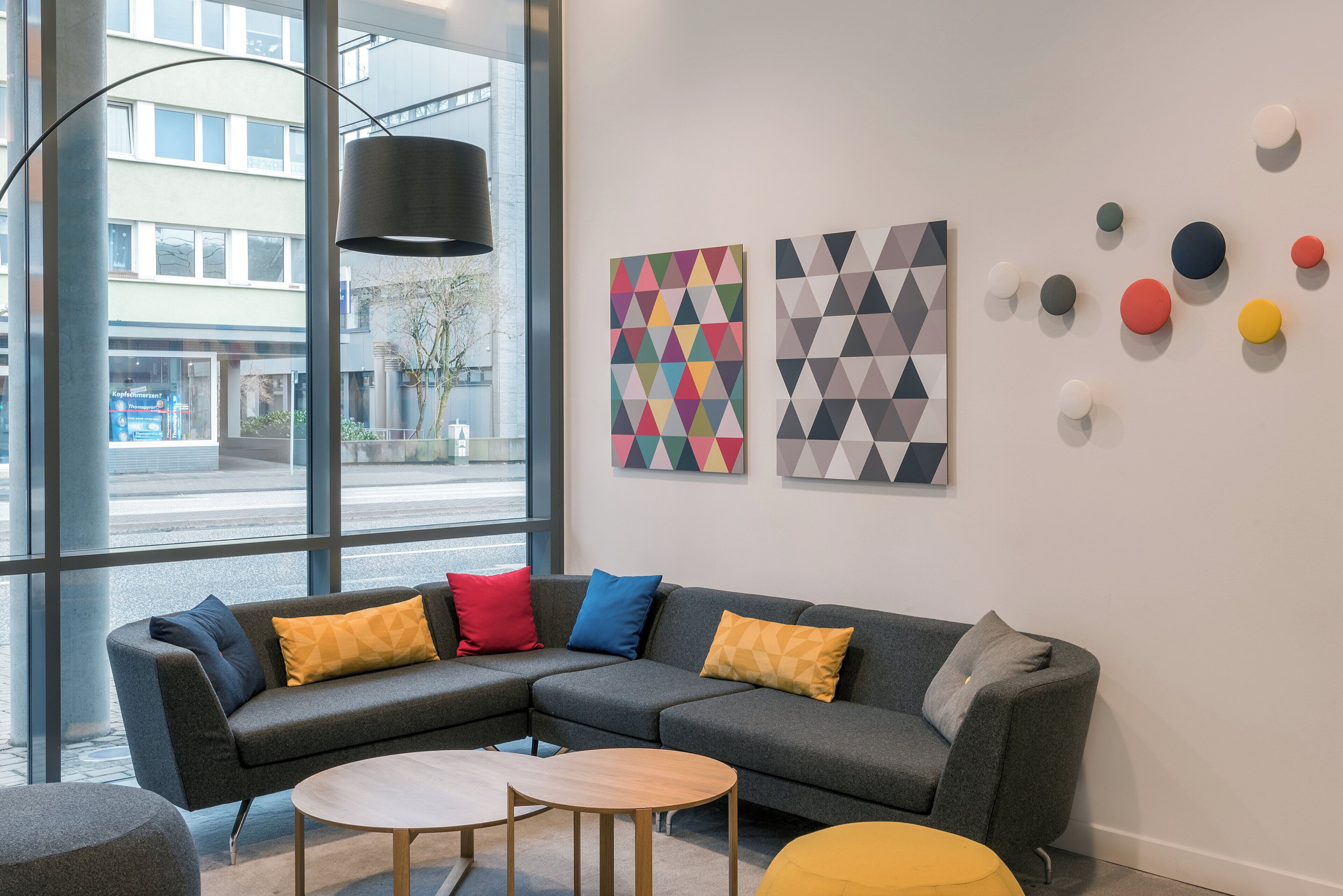 A modern interior with a grey sectional sofa, a round wooden table, and colorful geometric wall art.