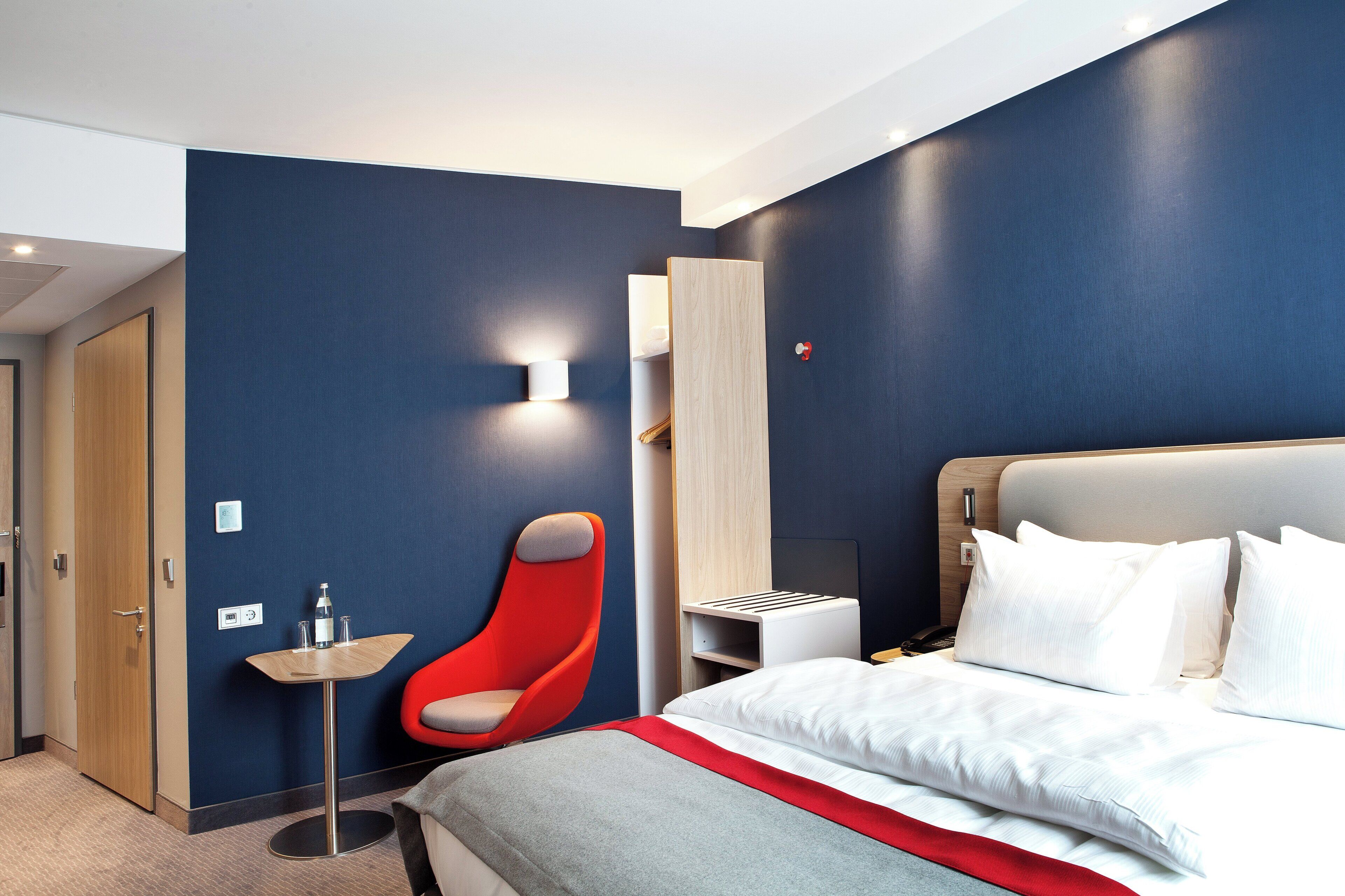 Quarto de hotel moderno com cama, uma cadeira vermelha, uma mesa pequena e uma luminária fixada na parede.