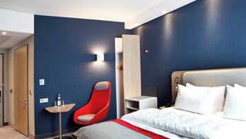Quarto de hotel moderno com cama, uma cadeira vermelha, uma mesa pequena e uma luminária fixada na parede.