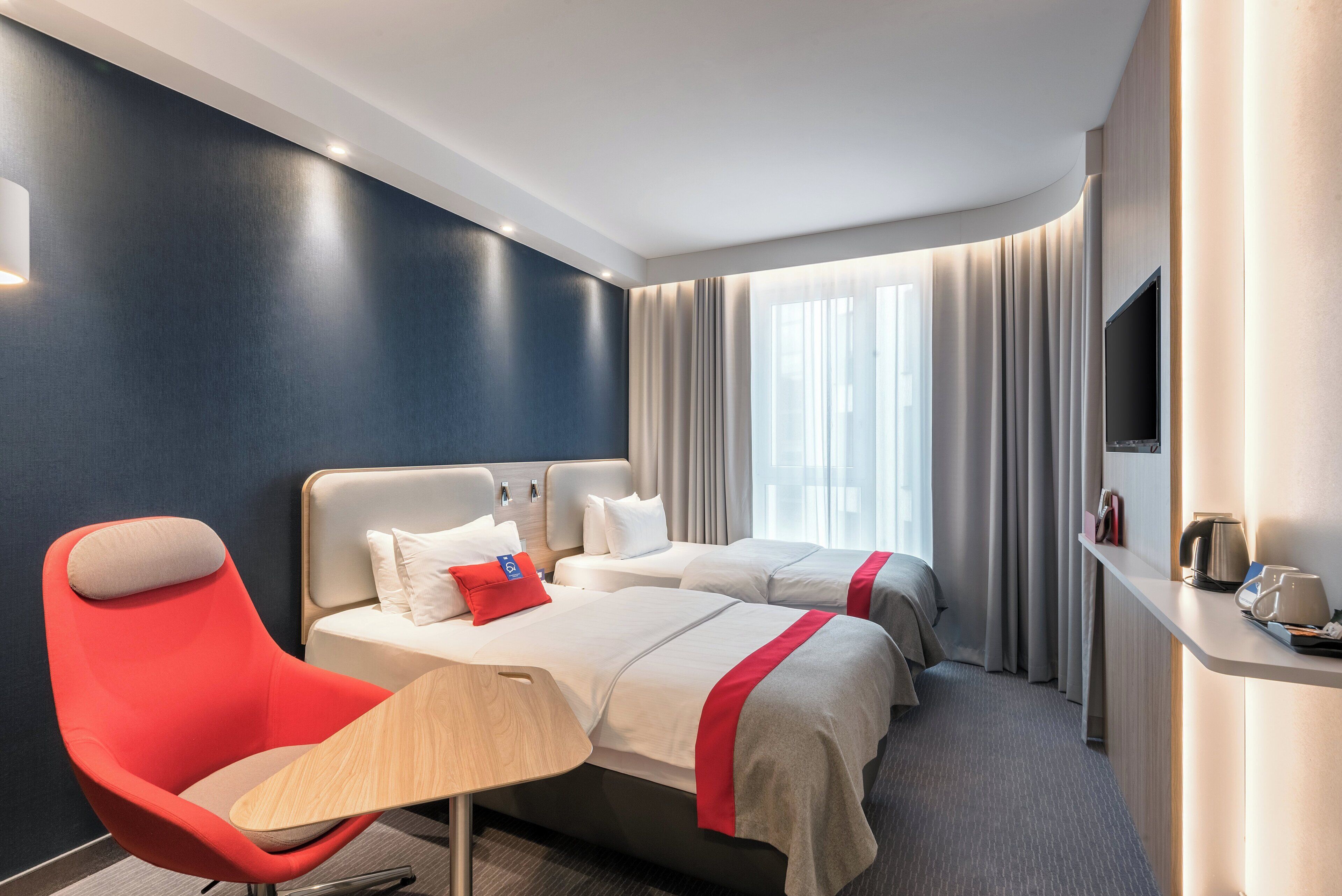 Habitación de hotel con dos camas, una silla roja, un televisor de pantalla plana y una cafetera.