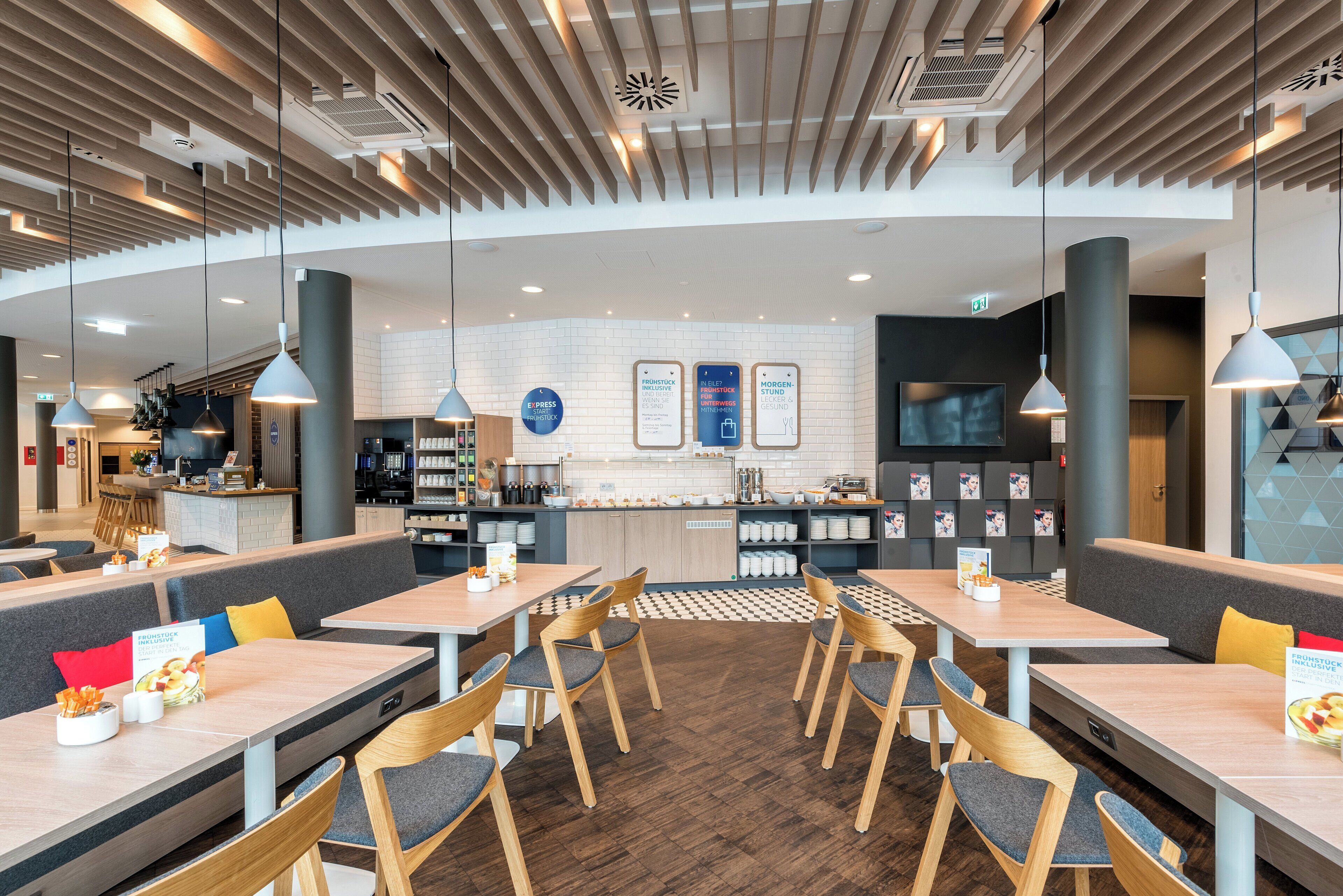 Interior moderno de restaurante con mesas de madera, asientos con cojines grises y una barra central con una pantalla de menú.