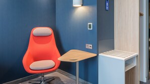 Una habitación de hotel moderna con una pared azul de acento, una silla roja, una pequeña mesa de madera y una lámpara fijada a la pared.